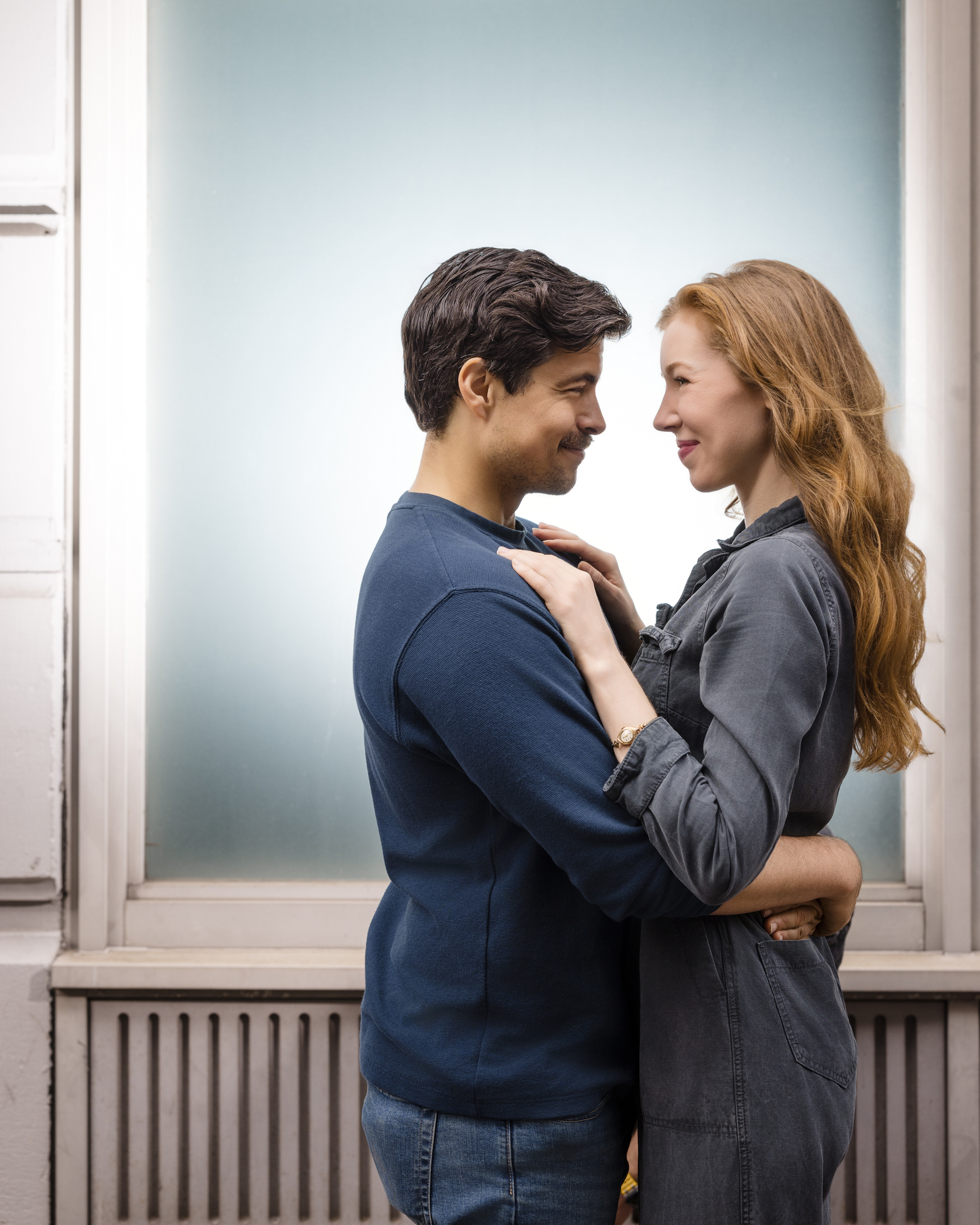 A man and woman stand facing each other indoors, smiling and holding each other’s arms in front of a frosted window during an NYC photo walk.