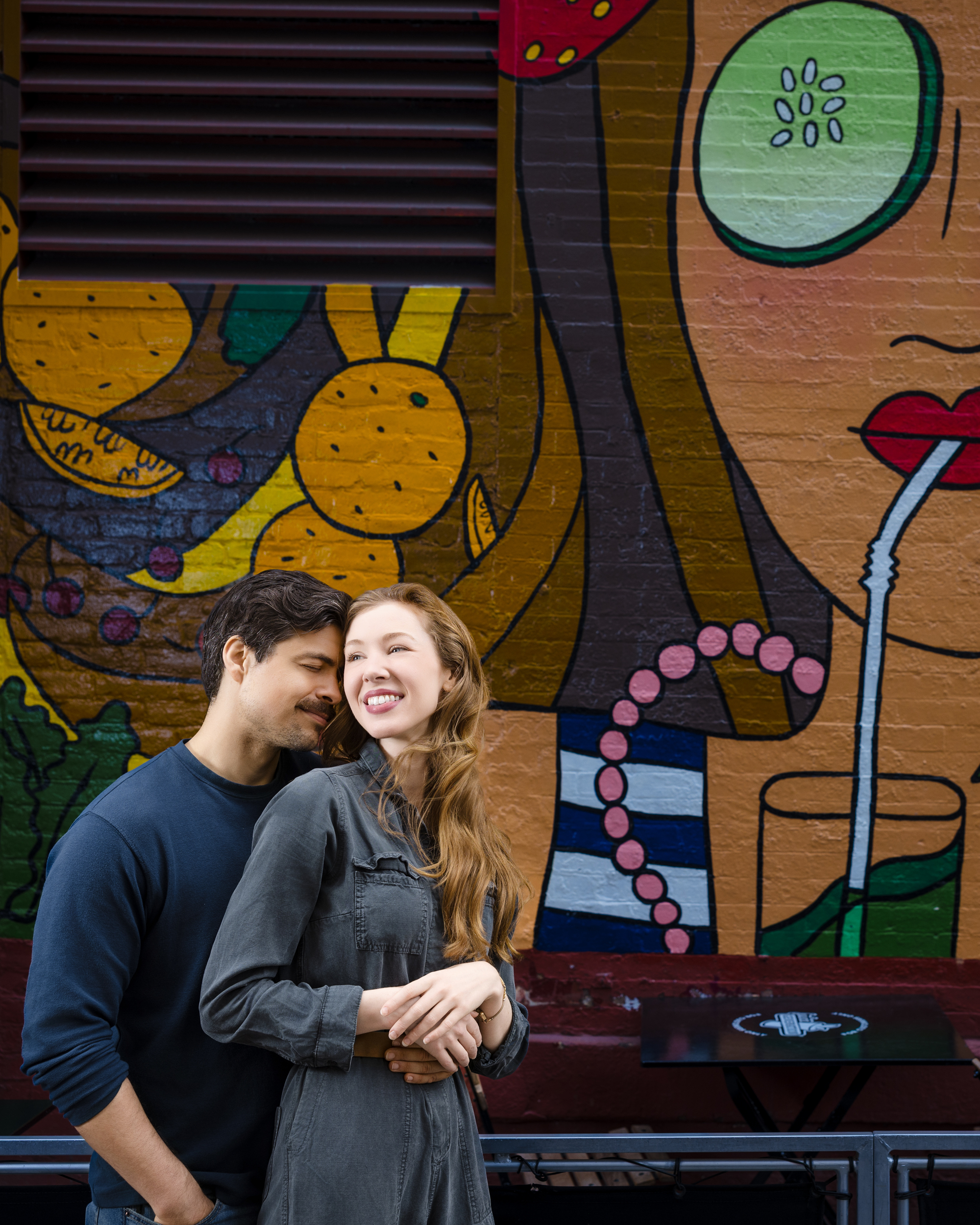 A man and woman stand in front of a colorful mural featuring a woman's face drinking through a straw. The woman smiles while the man leans in close behind her, capturing a candid moment during their NYC photo walk.