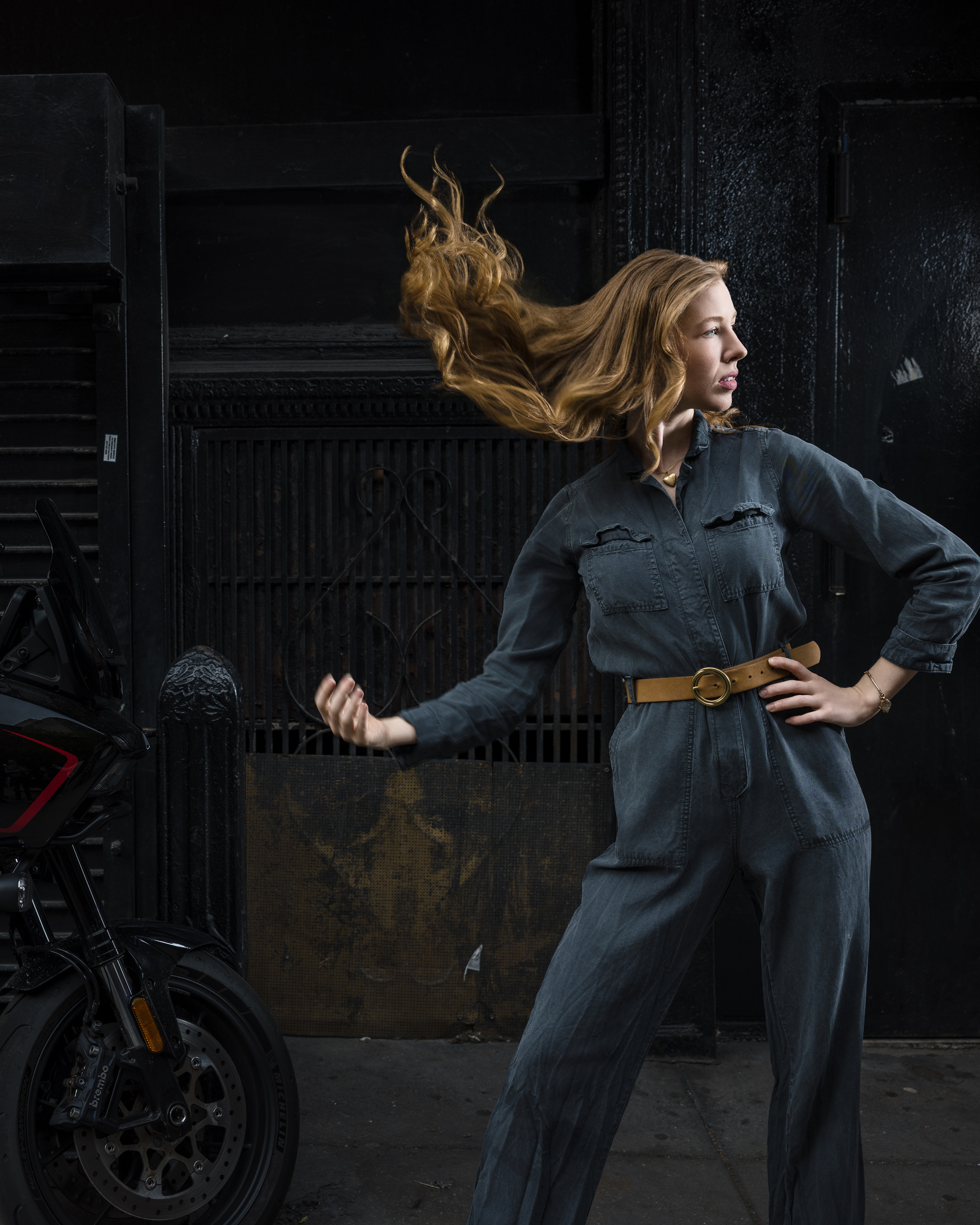 A woman in a gray jumpsuit stands next to a motorcycle during an NYC photo walk, her hair flipping in the air and one arm bent at her waist.