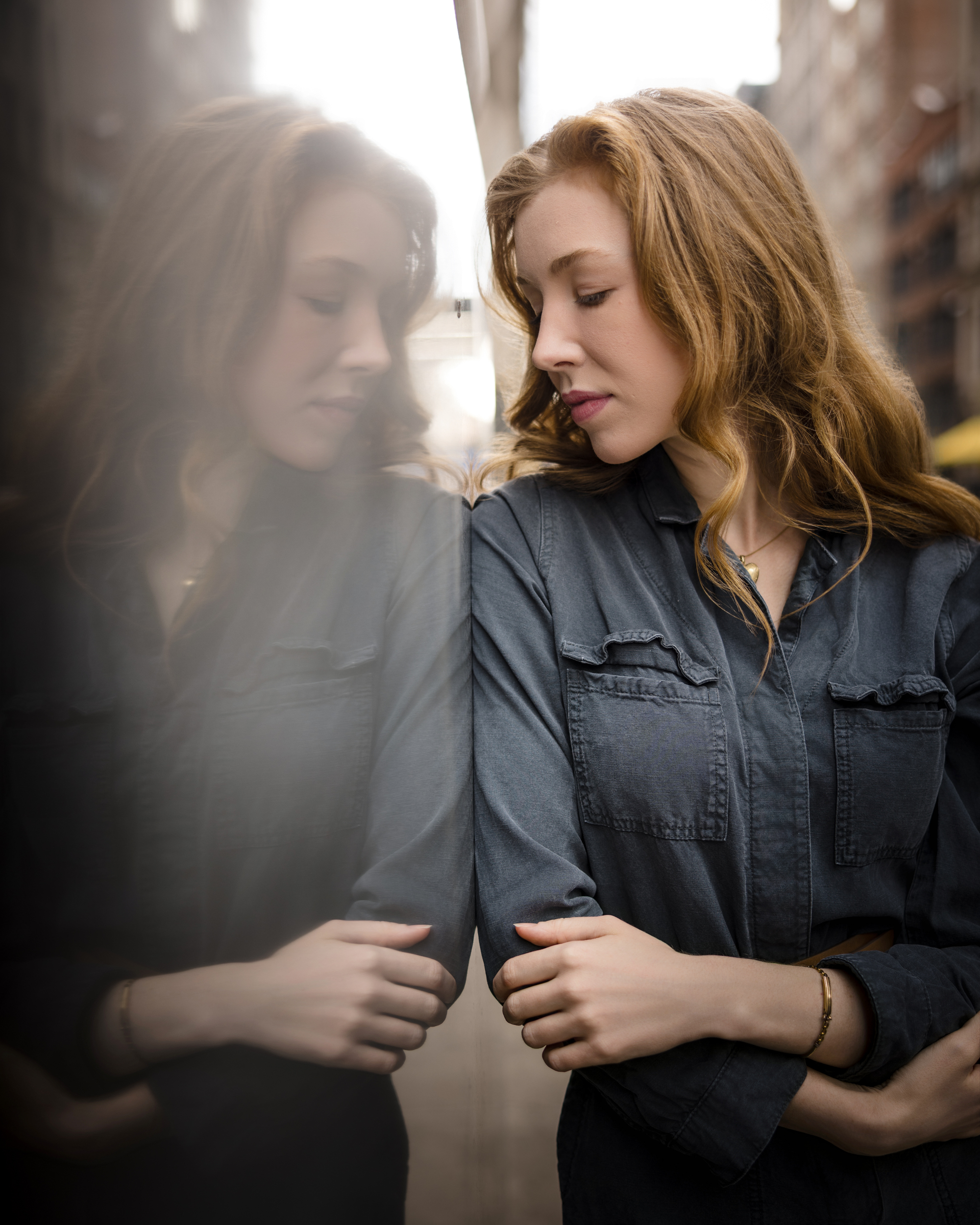 A woman with long hair in a dark shirt leans against a reflective glass surface during an NYC photo walk, looking down with her arms crossed as her reflection appears beside her.