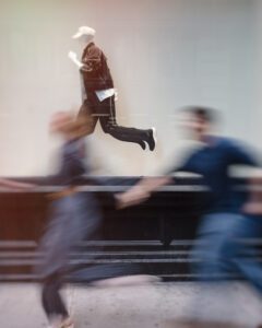 A mannequin in a running pose is displayed behind glass, while two blurred figures dash past on the sidewalk in the foreground during an NYC photo walk.