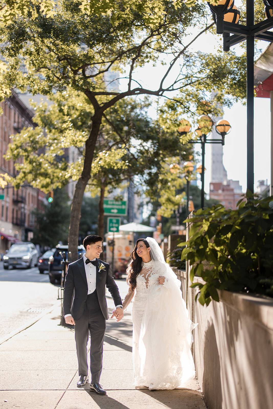 A bride and groom walk hand in hand on a city sidewalk, dressed in formal wedding attire, with trees and buildings hinting at a romantic Guastavino's wedding in the background.