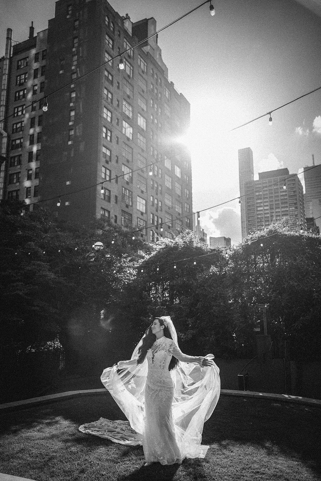 A bride in a wedding dress stands on a lawn with string lights overhead at a Guastavino's wedding, with tall buildings and sunlight visible in the background.