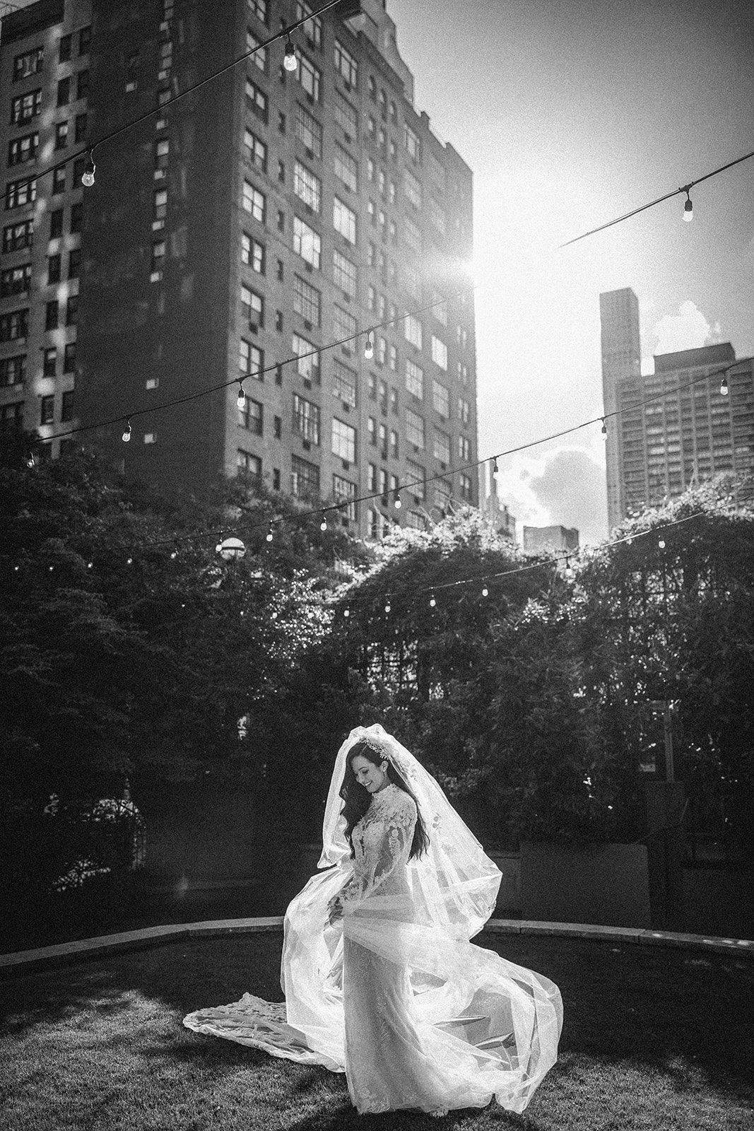 A bride in a long veil and gown stands on grass in an urban outdoor setting, with tall buildings and string lights twinkling—capturing the elegant charm of a Guastavino's wedding.