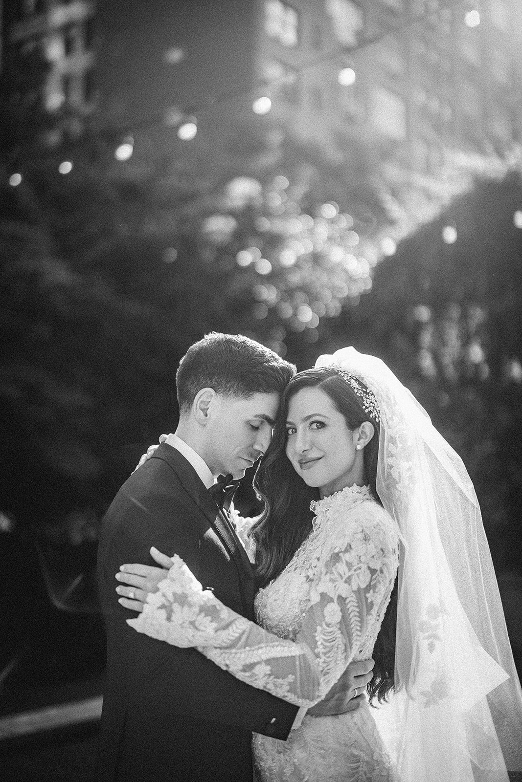 A bride and groom in formal wedding attire embrace outdoors at their Guastavino's wedding, with the bride looking at the camera and the groom resting his head on her. String lights and trees twinkle softly in the background.