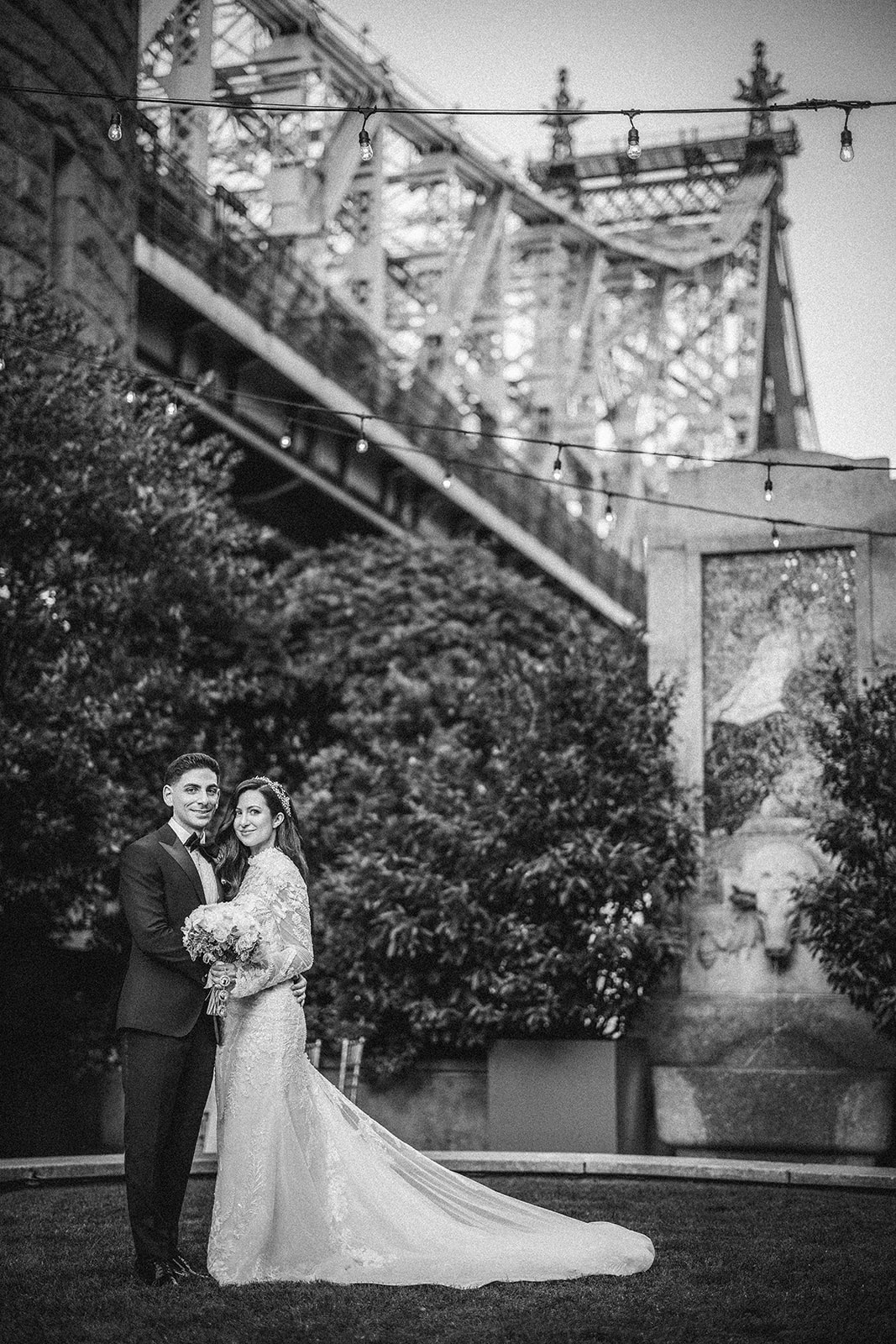 A bride and groom in formal attire pose together outdoors beneath a large bridge at a Guastavino's wedding, with string lights and greenery in the background.