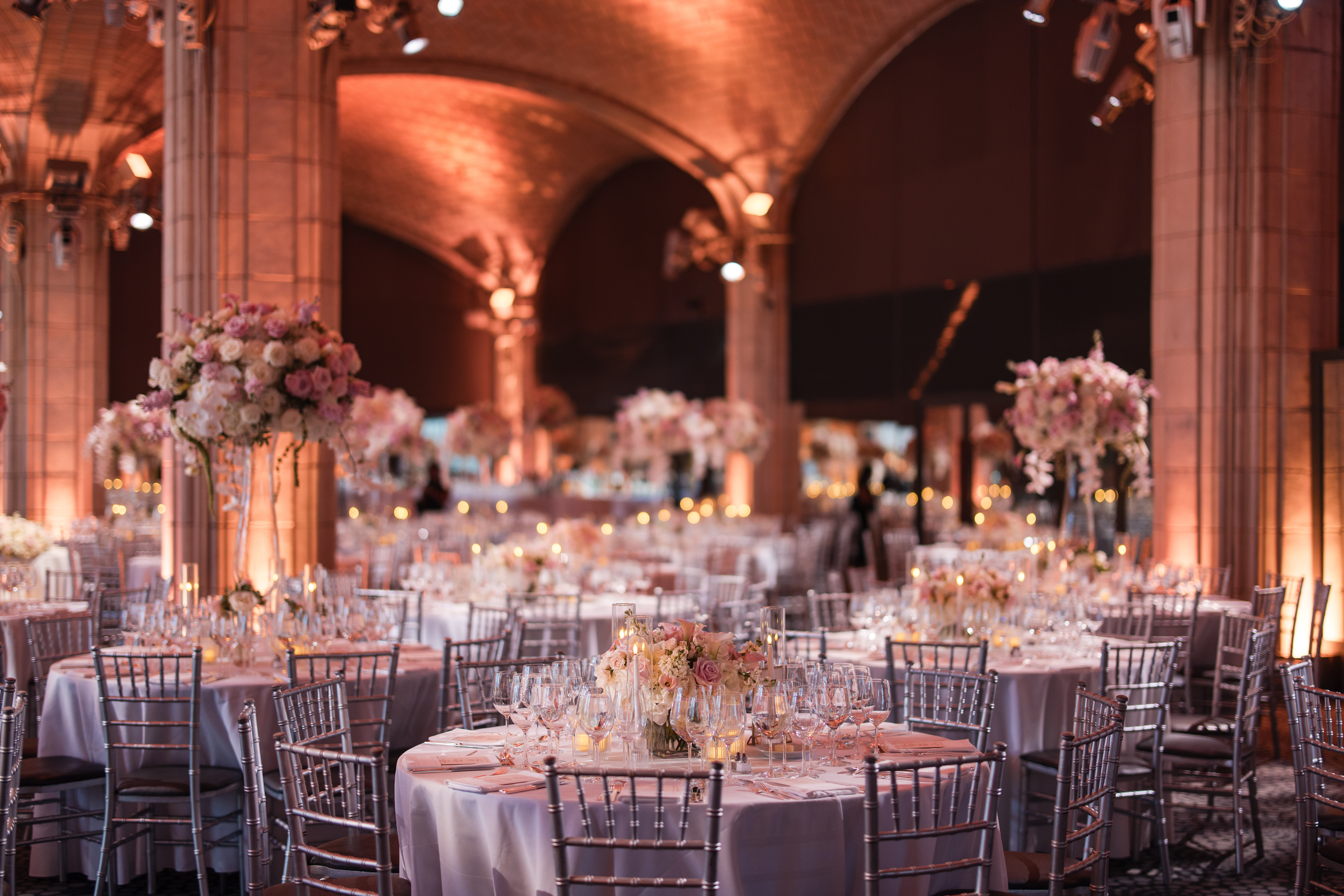 A formal event space with round tables set for dinner, silver chairs, floral centerpieces, and warm lighting under arched ceilings—perfect for a classic Guastavino's wedding.