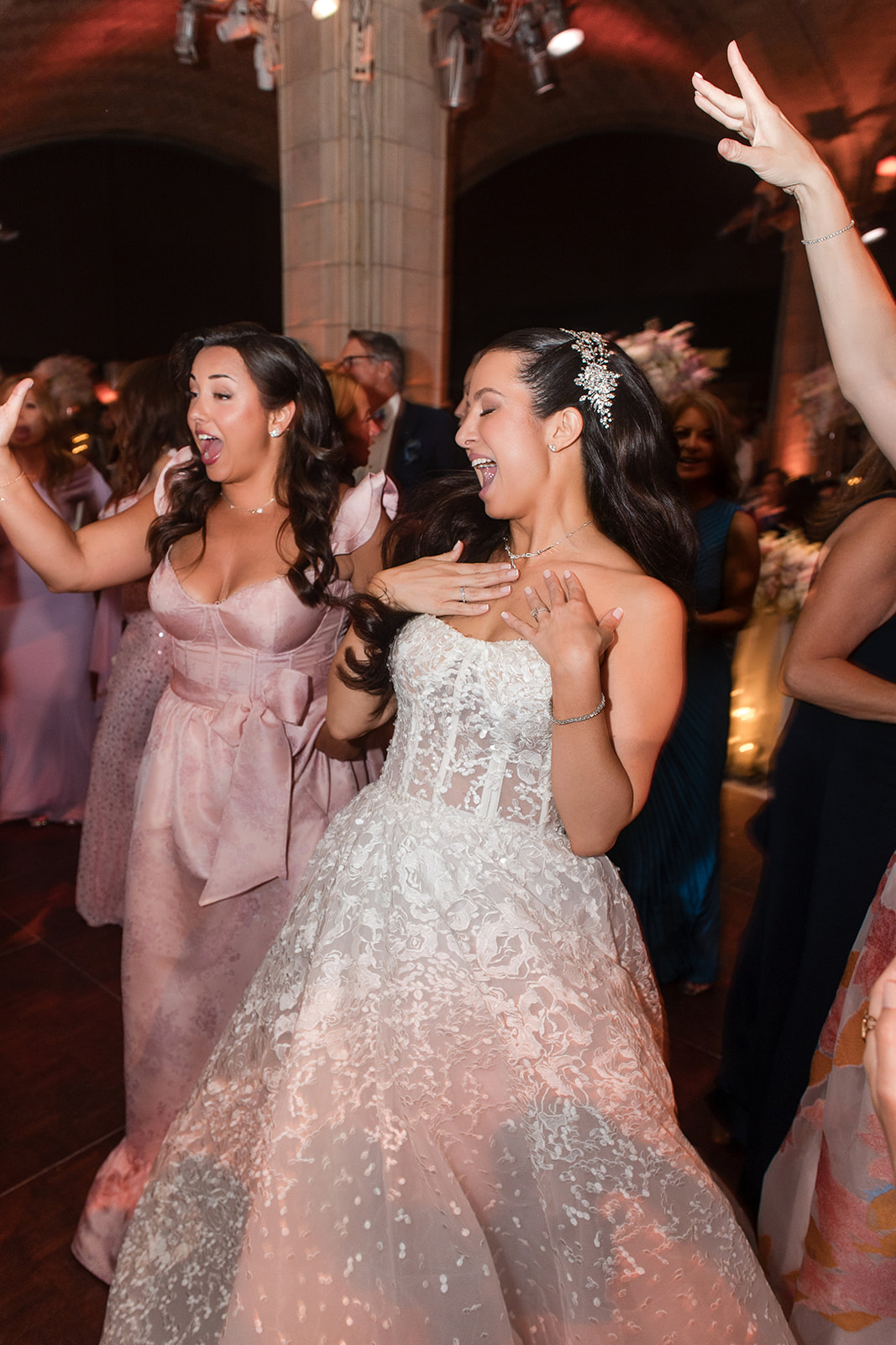Two women in formal dresses, one in a white wedding gown, smile and dance joyfully at a Guastavino's wedding, with other guests celebrating in the elegant indoor venue.