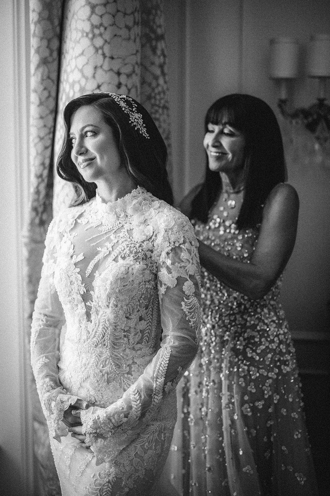 A bride in a lace wedding dress stands by a window, smiling, as another woman adjusts her hair. Both wear embellished dresses. The black and white scene captures the elegance of a Guastavino's wedding.