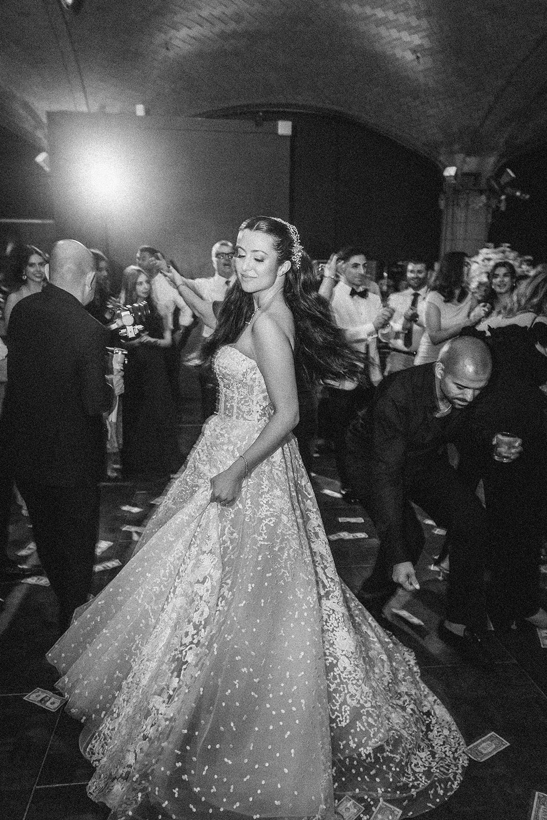 At a lively Guastavino's wedding, a woman in a formal gown dances the Greek money dance as a man nearby crouches to pick up money from the floor, with guests watching in the background.
