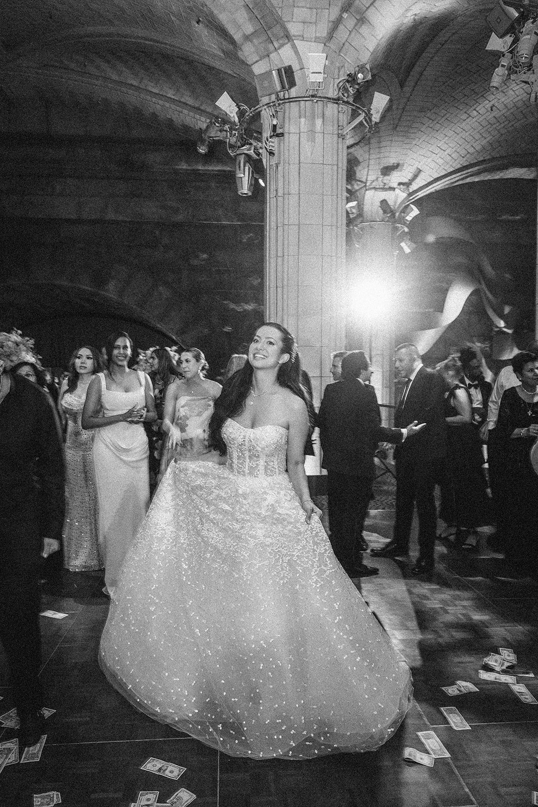 A woman in a sparkly gown smiles and holds her dress at a Guastavino's wedding, with guests nearby and money scattered on the floor after lively Greek dancing.
