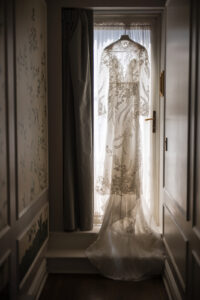 A sheer, long-sleeved wedding dress with floral lace detailing hangs on a door in front of a window, softly lit by natural light—perfect for an elegant Guastavino's wedding.