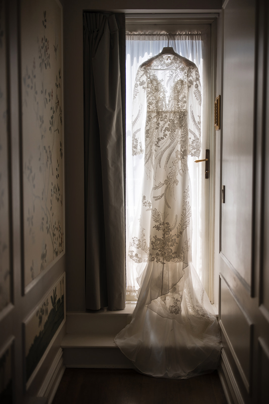 A sheer, long-sleeved wedding dress with floral lace detailing hangs on a door in front of a window, softly lit by natural light—perfect for an elegant Guastavino's wedding.