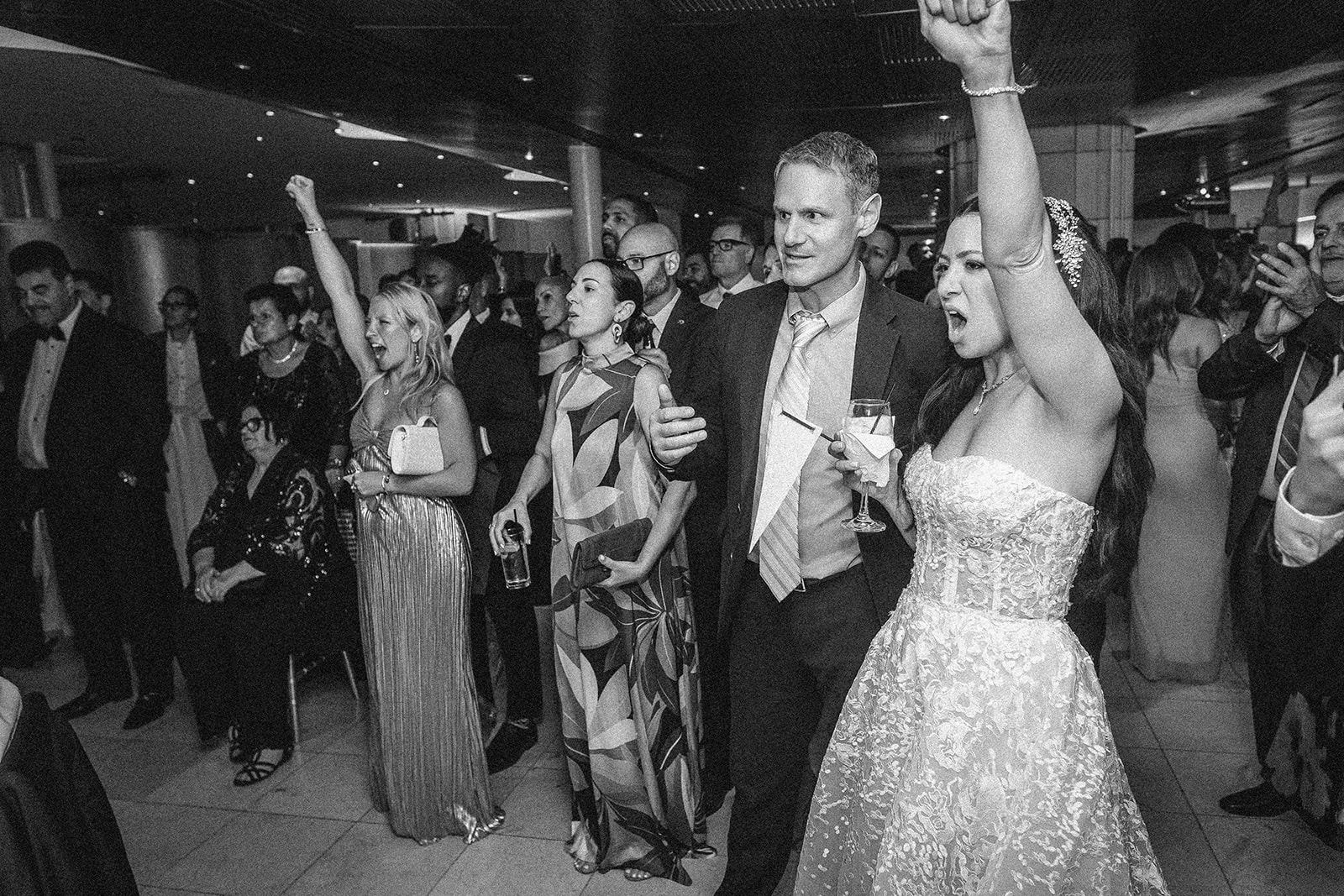 A group of formally dressed people, including a woman in a wedding gown, celebrate indoors with raised arms and drinks at what appears to be a Guastavino's wedding afterparty, watching the NY Knicks win.
