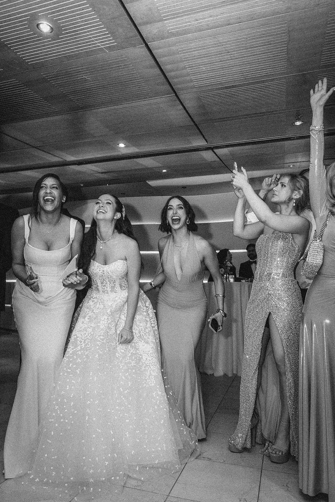 A group of women in formal dresses laugh and gesture excitedly at an indoor event, possibly watching the Knicks win at a Guastavino's wedding afterparty.