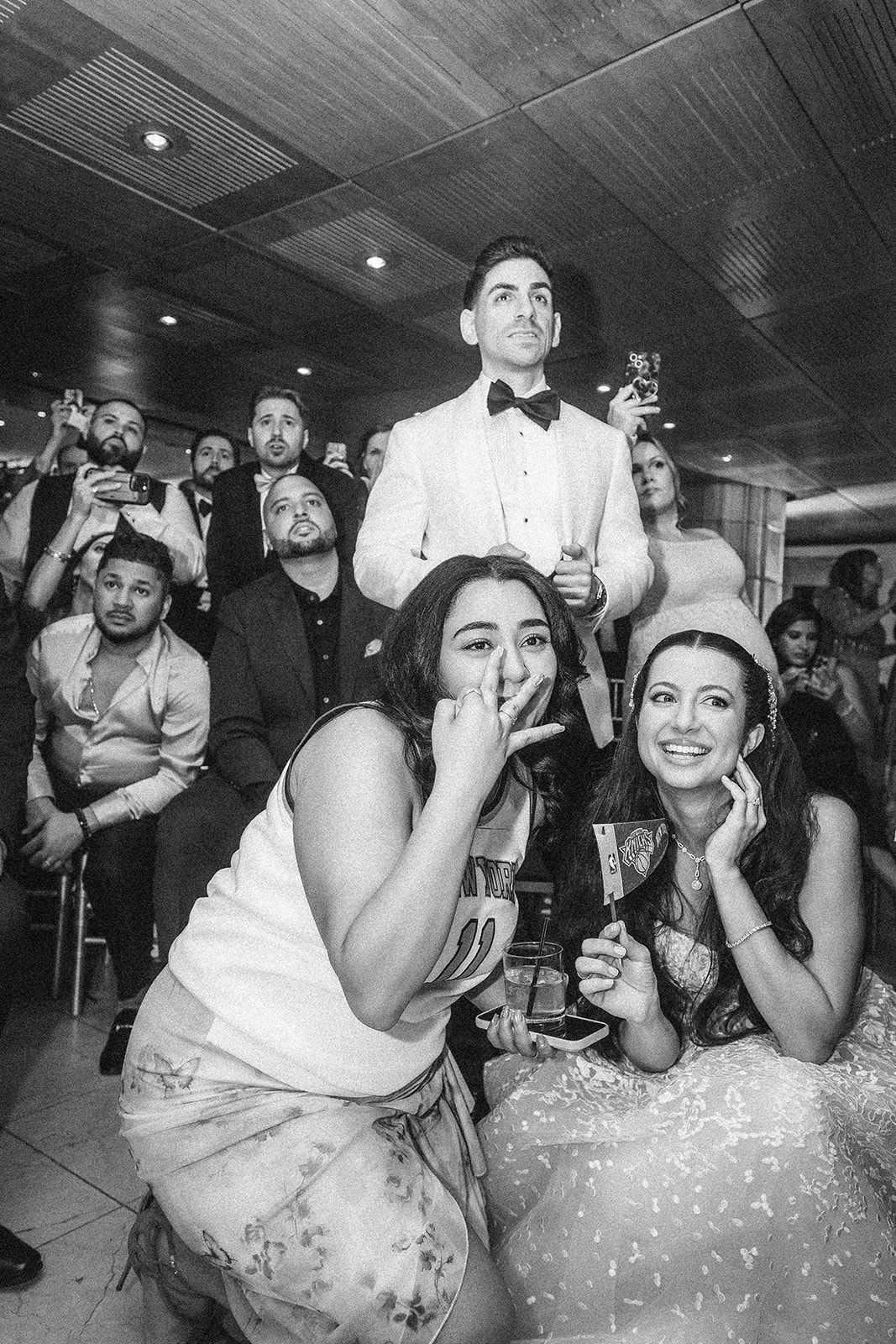 A group of people at an indoor event, possibly a Guastavino's wedding, with a woman in a dress and another in a tank top kneeling and smiling in the foreground, while others watch and take photos—maybe even watching the Knicks win.