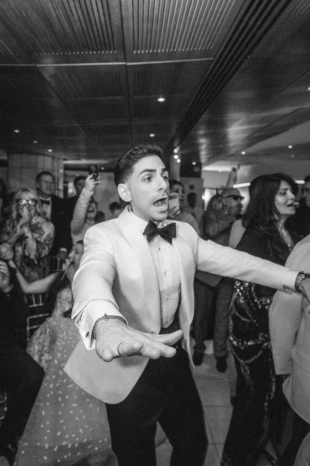 A man in a white tuxedo and bow tie gestures with outstretched arms at a Guastavino's wedding, surrounded by people who are celebrating the Knicks in five.