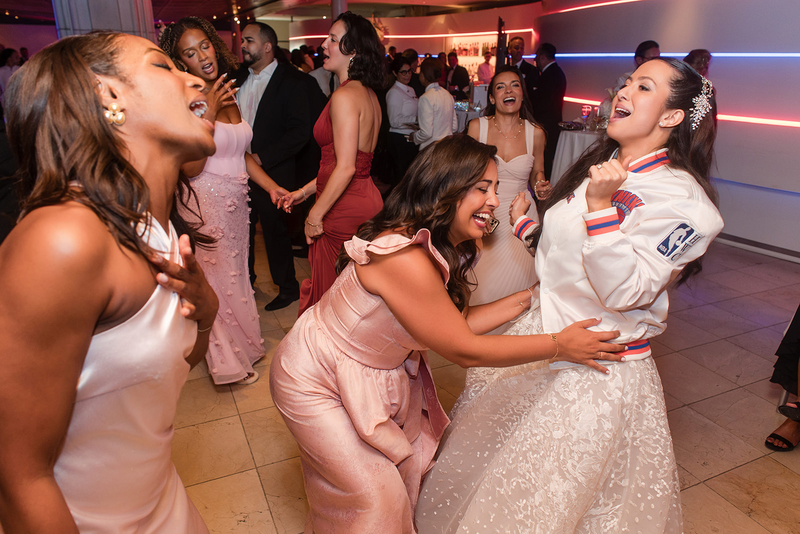 A group of women in formal attire laughing and dancing together at an indoor event with colorful lighting, celebrating a Knicks win at Guastavino's wedding.
