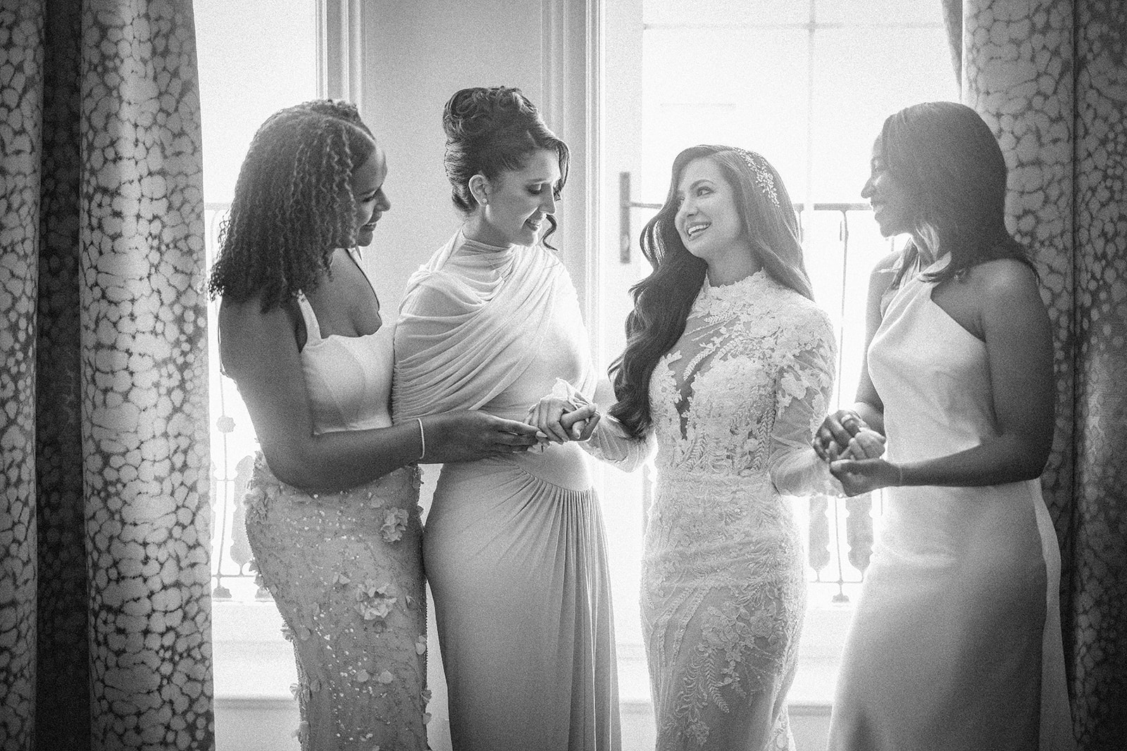Four women in elegant dresses stand together indoors at a Guastavino's wedding, smiling and holding hands, with light streaming through a window behind them.