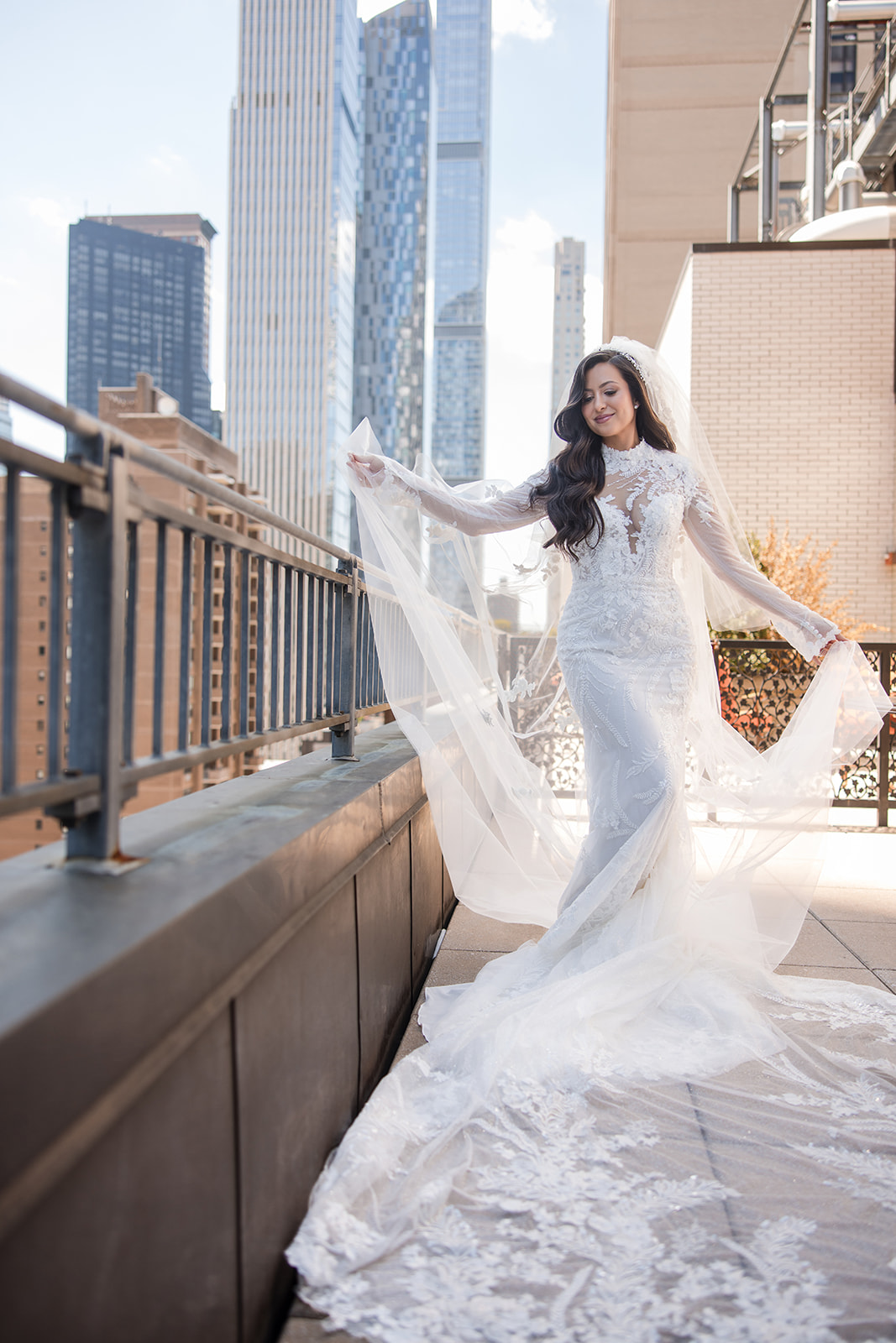 A woman in a white lace wedding dress and veil stands on a city rooftop, holding out her veil with skyscrapers visible in the background, capturing the elegant spirit of a Guastavino's wedding.