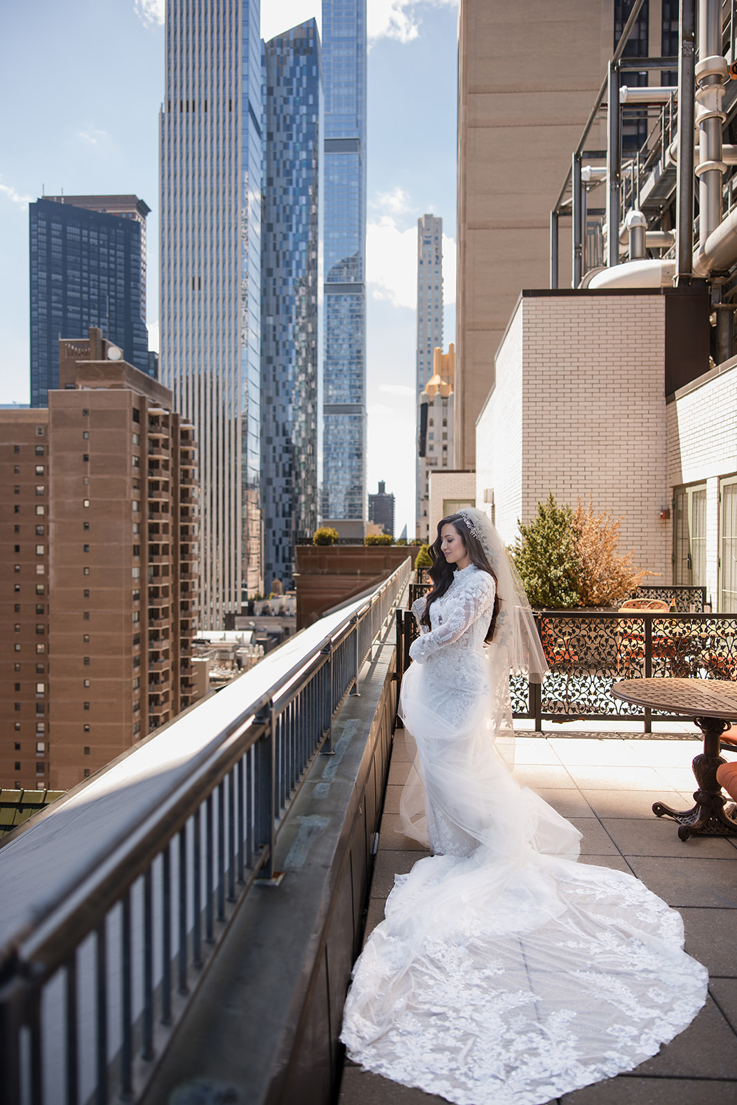 A bride in a white lace gown and veil stands on a city rooftop balcony with tall buildings in the background, capturing the elegance of a Guastavino's wedding.