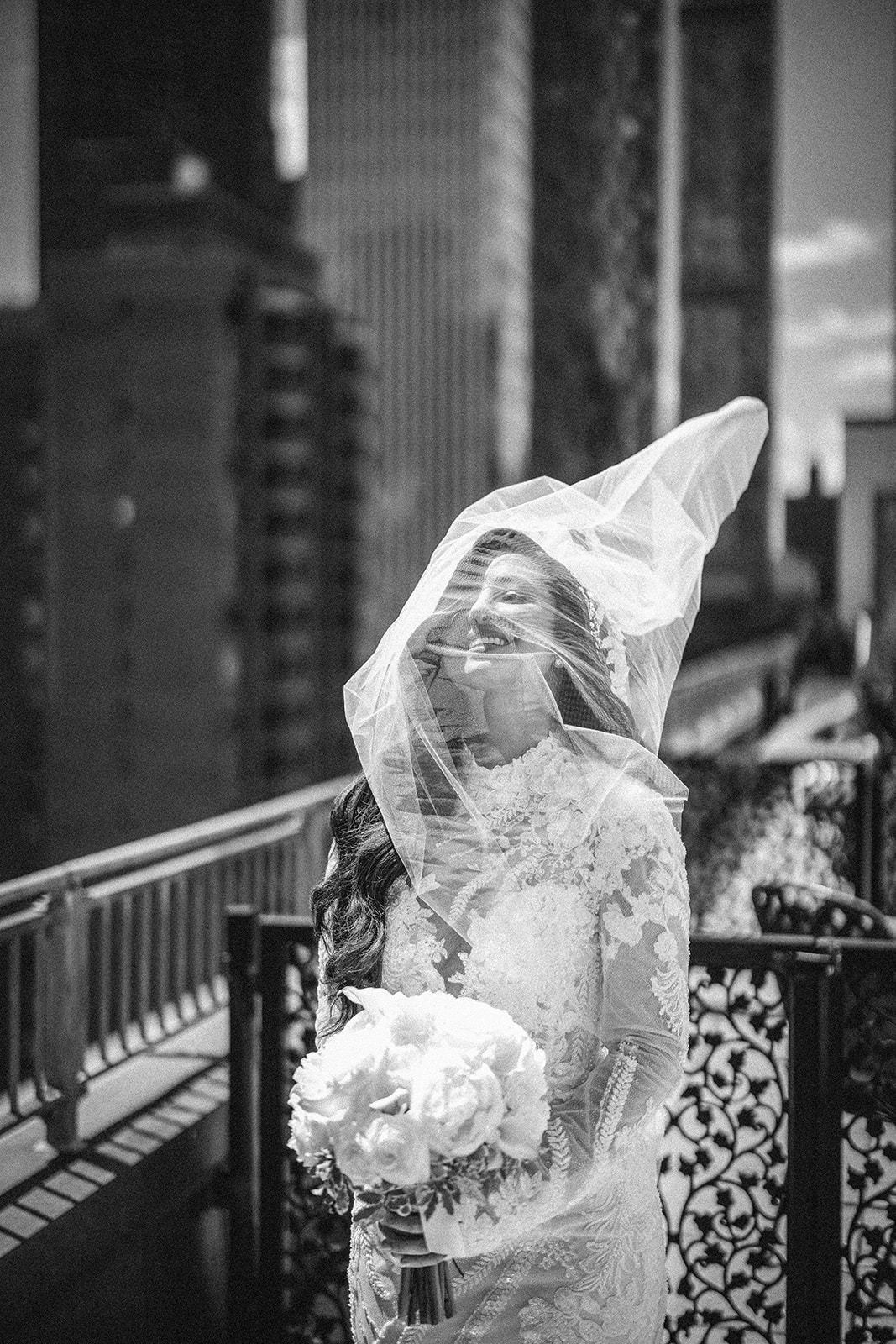 A bride in a lace wedding dress holds a bouquet and stands on a balcony at her Guastavino's wedding, with her veil blowing across her face and city buildings in the background.