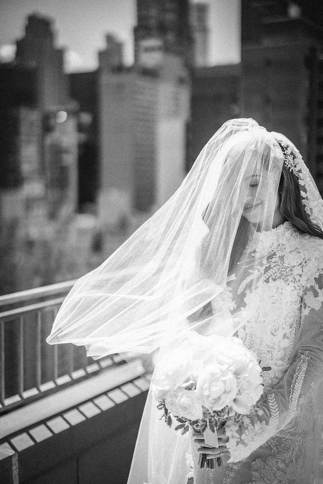 A bride in a lace gown and veil holds a bouquet of flowers while standing outdoors on a balcony, the blurred city skyline behind her setting the perfect scene for a memorable Guastavino's wedding.