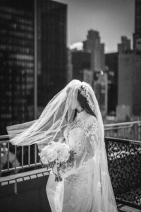 A bride in a lace wedding dress and veil holds a bouquet of flowers while standing on a city rooftop, evoking the elegance of a Guastavino's wedding with tall buildings in the background.