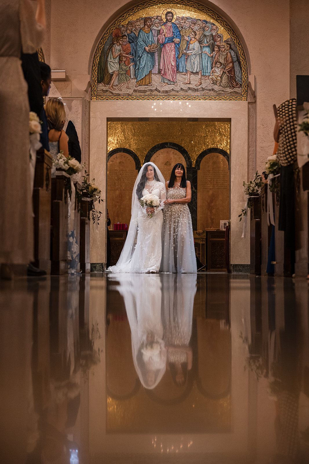 A bride in a white dress and veil stands with a woman in a silver gown at the entrance of a church, with their reflections shimmering on the polished floor—capturing the elegance of a Guastavino's wedding beneath the mosaic above.
