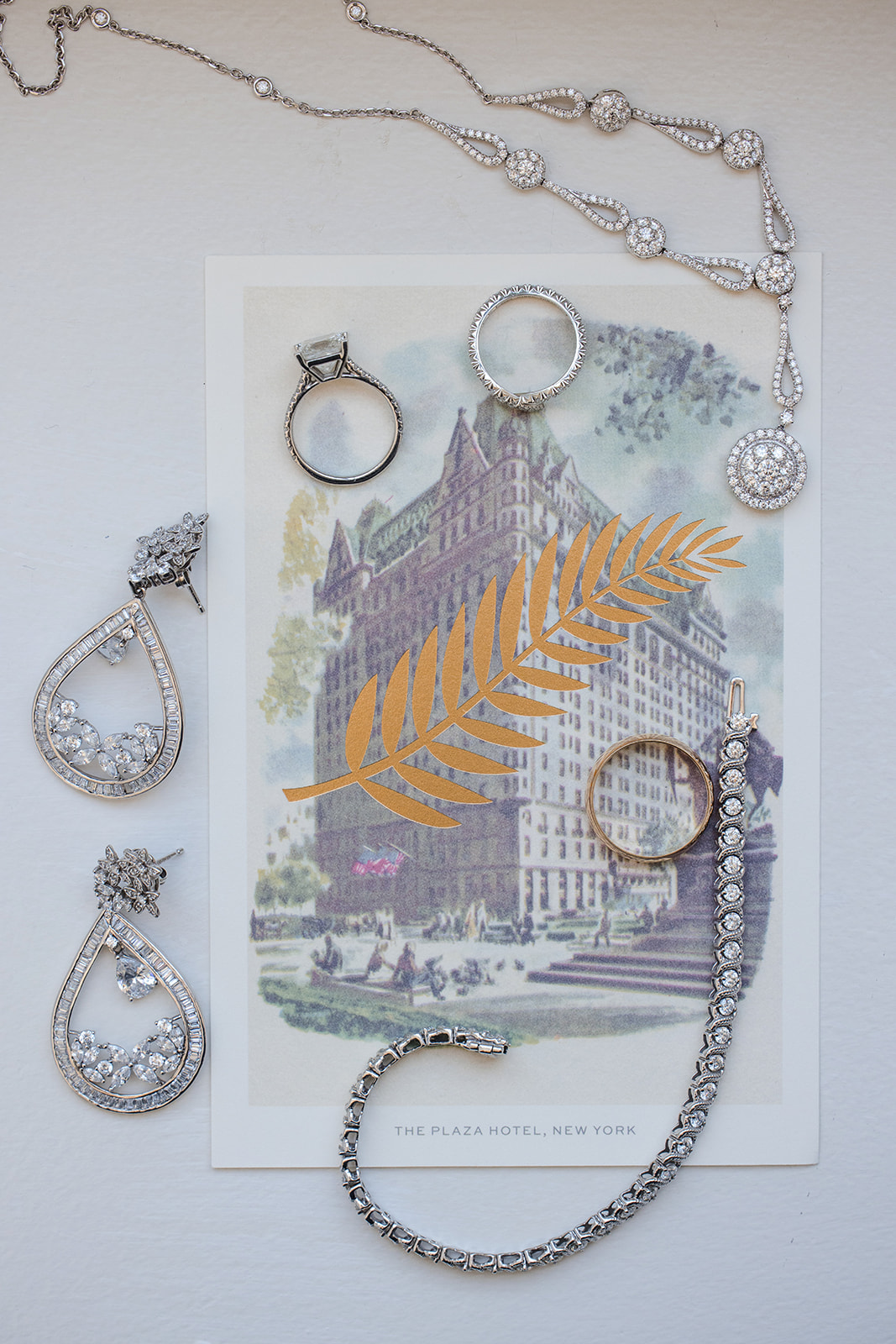Assorted diamond jewelry, including rings, earrings, a bracelet, and a necklace, arranged on a postcard featuring The Plaza Hotel, New York with a gold leaf design—perfect for a Guastavino's wedding celebration.