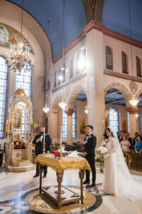 Bride and groom stand beside a decorated table during a Guastavino's wedding ceremony in an ornate church, with guests seated and religious icons visible in the background.