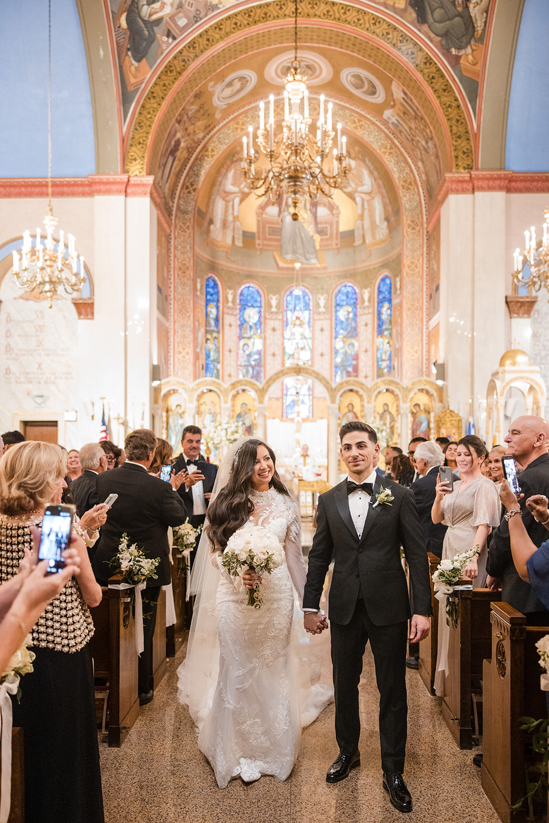 A bride and groom walk down the aisle together in a decorated church as guests celebrate their Guastavino's wedding and snap joyful photos.