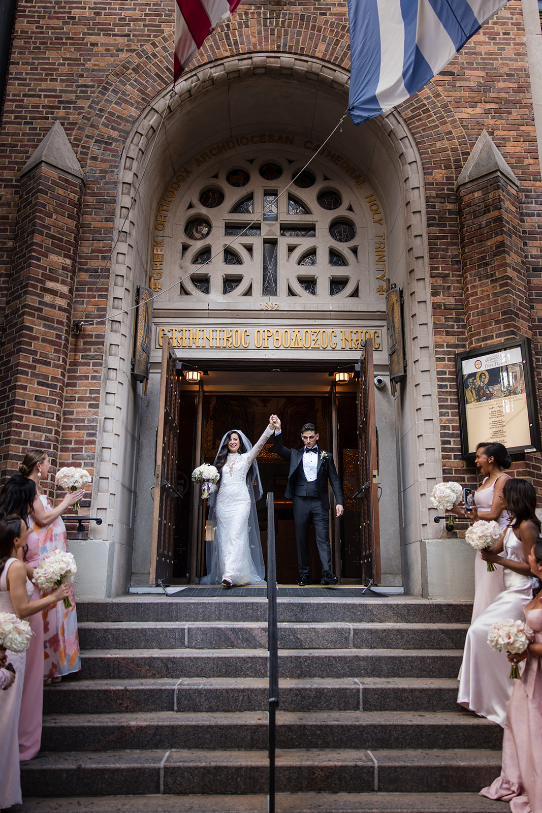 A bride and groom exit a church holding hands, smiling, as bridesmaids in pink dresses stand on the steps holding bouquets—capturing the joy of a Guastavino's wedding celebration.