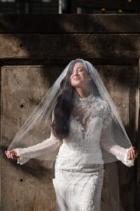 A woman in a white lace wedding dress and veil stands outside against a concrete wall, eyes closed, holding the sides of her veil—a serene moment before her Guastavino's wedding.