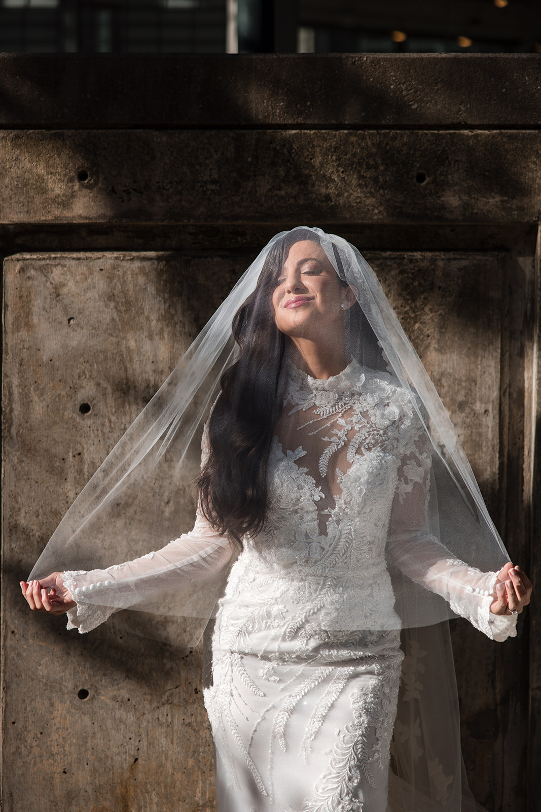 A woman in a white lace wedding dress and veil stands outside against a concrete wall, eyes closed, holding the sides of her veil—a serene moment before her Guastavino's wedding.