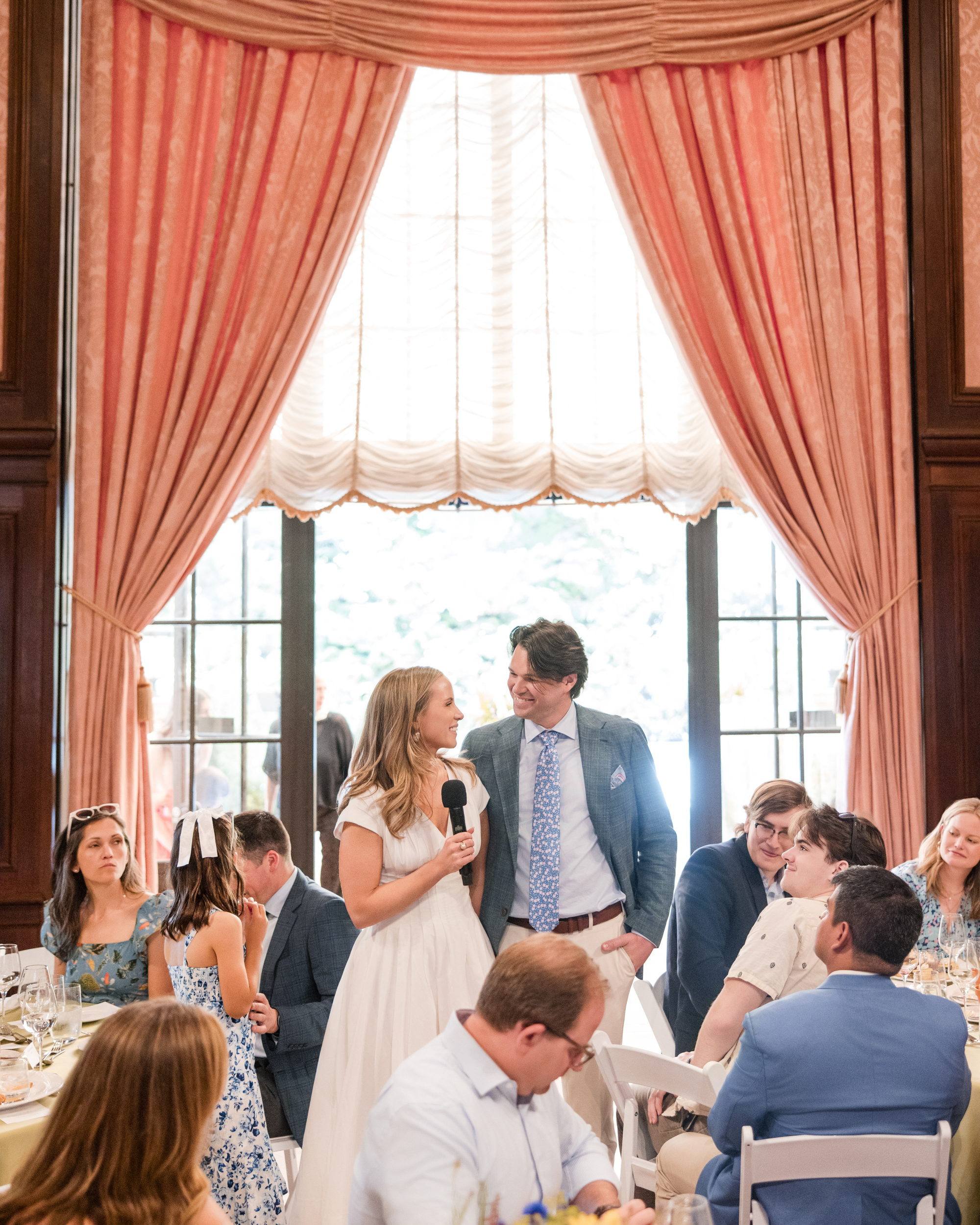 At a Longwood Gardens welcome party, a woman in a white dress speaks into a microphone beside a man in a suit, surrounded by seated guests and large draped windows in the elegant indoor venue.