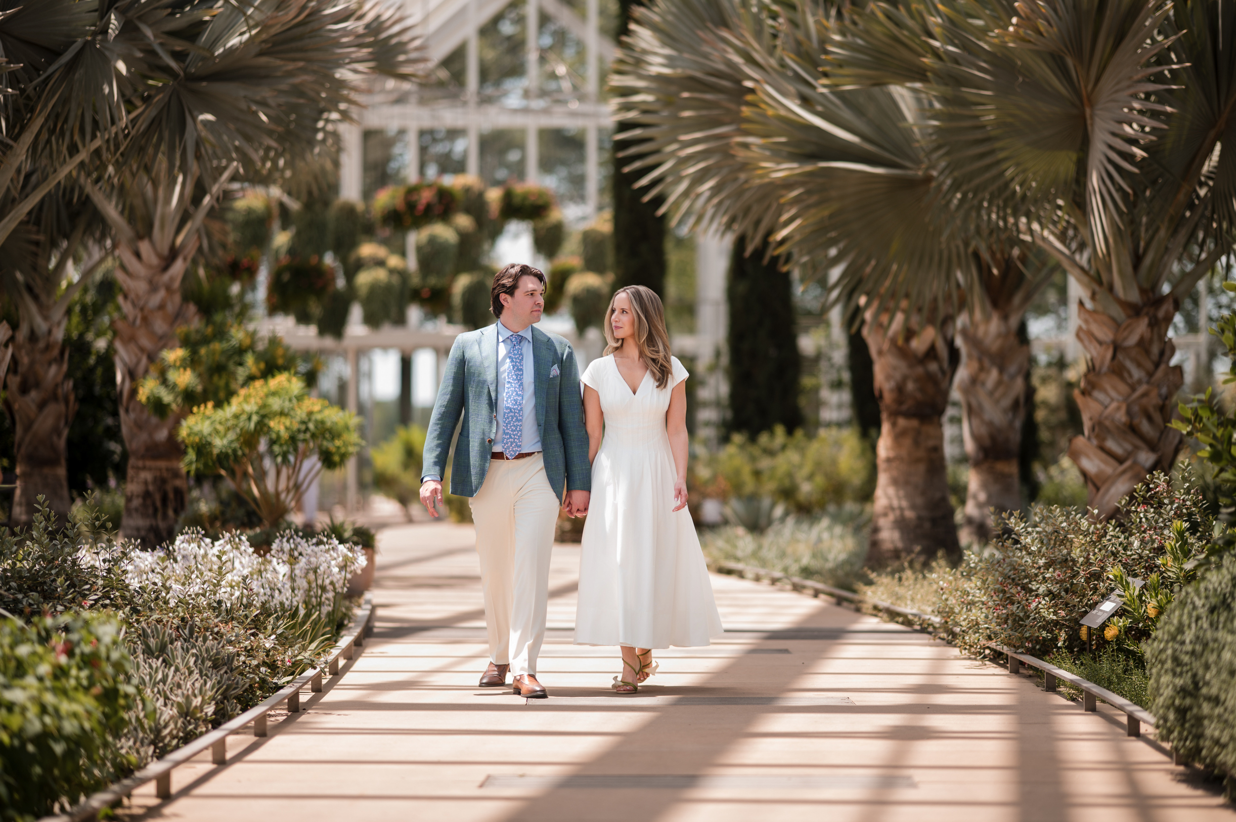 A man and woman in formal attire walk hand in hand on a garden pathway at a Longwood Gardens welcome party, surrounded by palm trees and lush greenery.