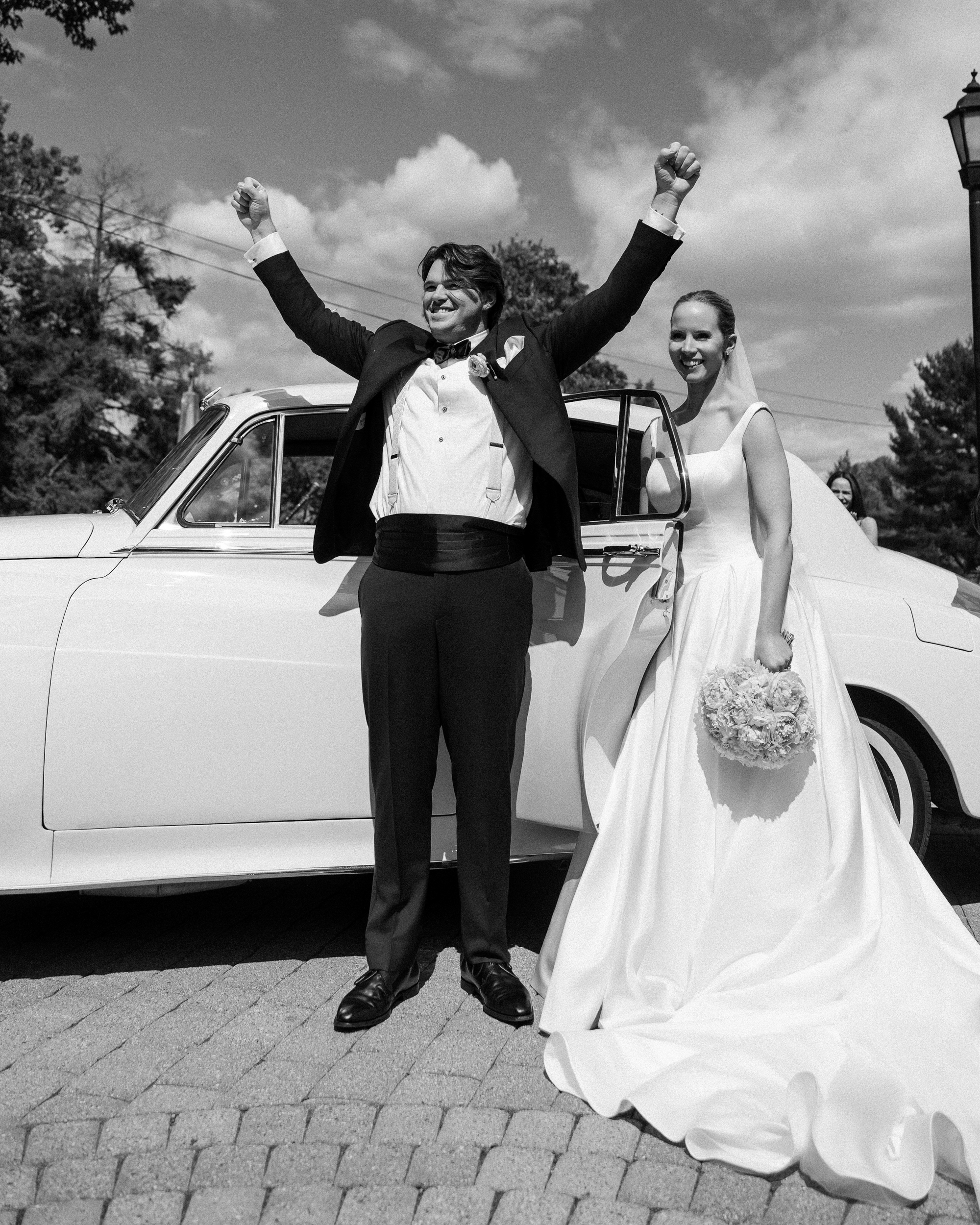 A bride and groom stand by a classic car at the Jasper Crane Rose Garden, the groom raising his arms and smiling, while the bride holds a bouquet and smiles at the camera—capturing timeless wedding photos in this picturesque setting.