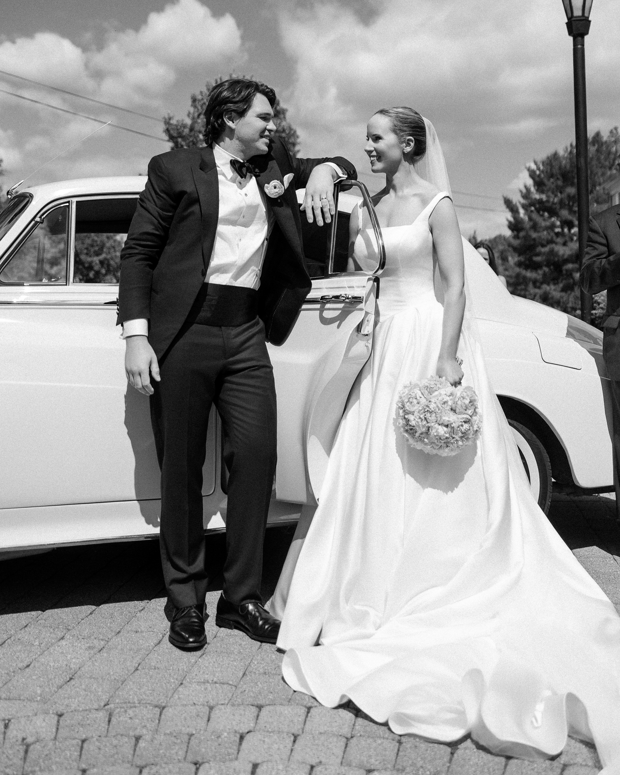Bride and groom stand beside a vintage car, smiling at each other in front of the Jasper Crane Rose Garden; the bride holds a bouquet and wears a long gown, while the groom is in a tuxedo—capturing timeless Jasper Crane Rose Garden wedding photos.