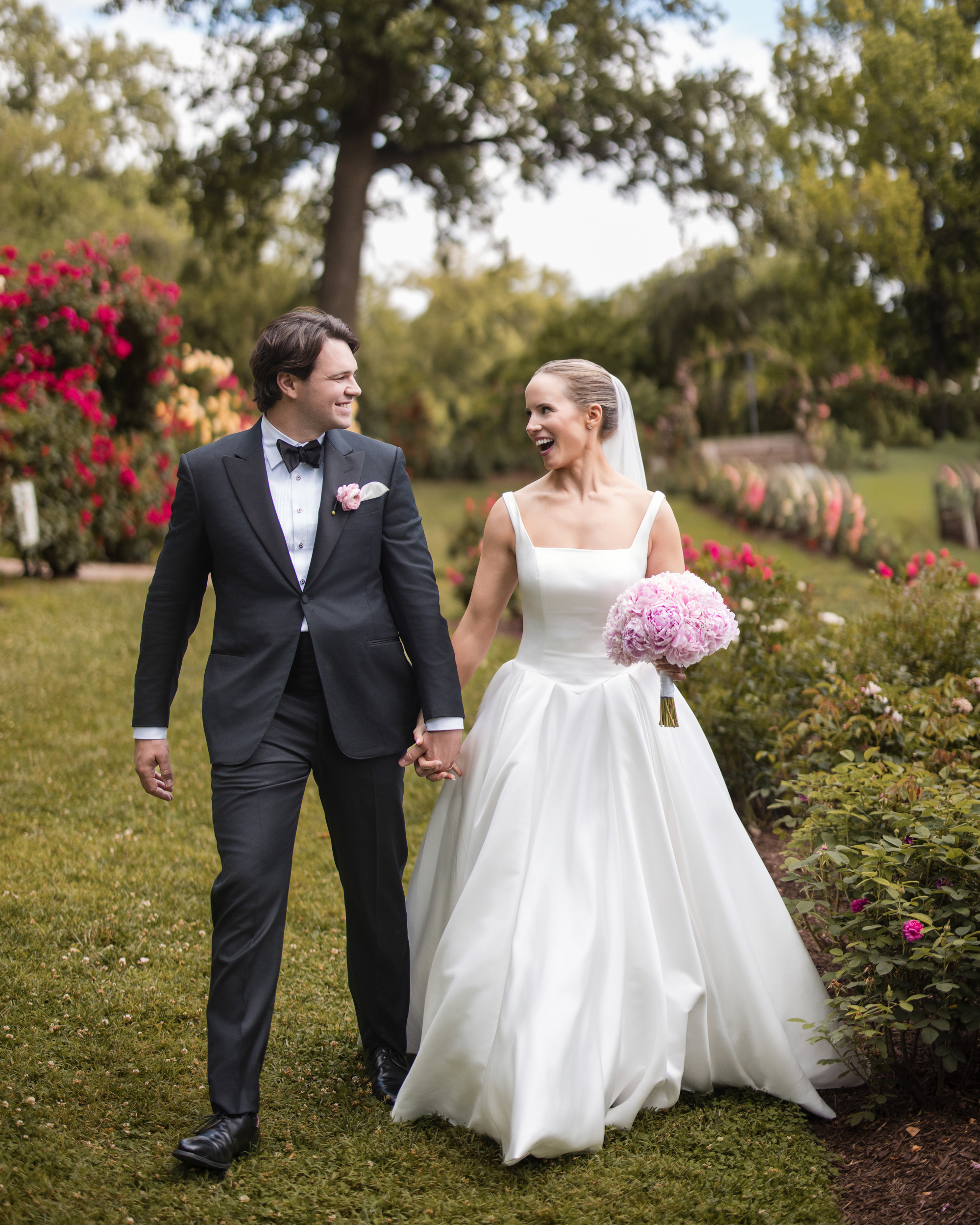 A bride in a white gown and a groom in a tuxedo hold hands and smile while walking outdoors in the Jasper Crane Rose Garden, surrounded by vibrant flowers—a perfect setting for unforgettable wedding photos.