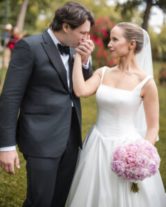 A groom in a black tuxedo kisses the bride's hand while she holds a pink bouquet. They stand outdoors at the Jasper Crane Rose Garden, both dressed in wedding attire, capturing stunning wedding photos.