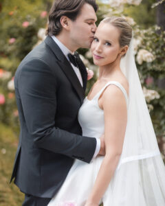 A groom in a black suit kisses a bride in a white dress and veil on the forehead as they pose outdoors among flowers, capturing stunning Jasper Crane Rose Garden wedding photos.