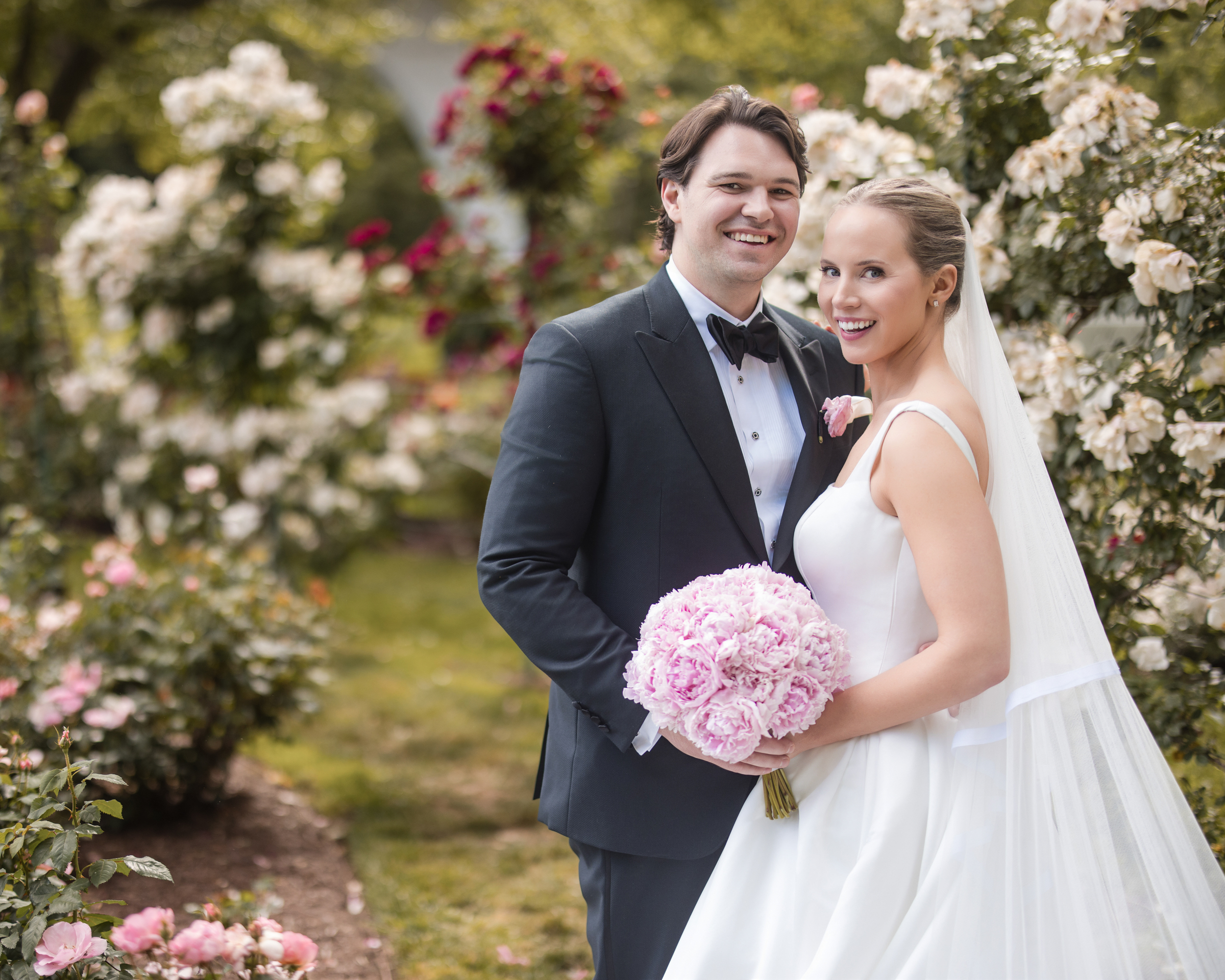 A bride in a white dress and groom in a tuxedo stand together in the Jasper Crane Rose Garden with blooming roses, both smiling at the camera. The bride holds a bouquet of pink flowers, creating timeless wedding photos amidst the floral beauty. A bride in a white dress and groom in a tuxedo stand together in the Jasper Crane Rose Garden with blooming roses, both smiling at the camera. The bride holds a bouquet of pink flowers, creating timeless wedding photos amidst the floral beauty.