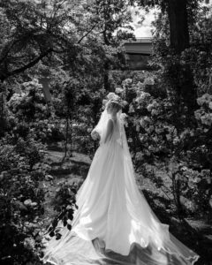 A bride in a long white gown and veil stands in the Jasper Crane Rose Garden, surrounded by blooming flowers and trees, with sunlight streaming through the foliage—creating perfect wedding photos.