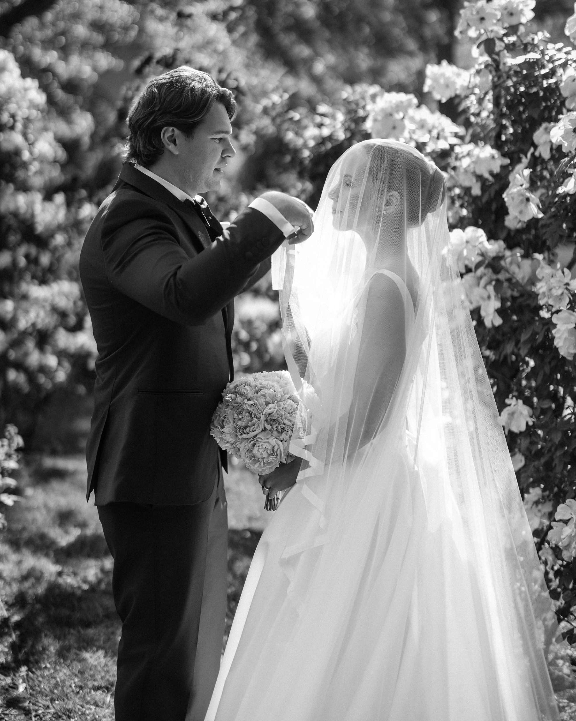 A groom lifts the bride’s veil as they stand facing each other in a garden, surrounded by blooming flowers. The bride holds a bouquet and wears a long veil and gown. Captured in black and white, this timeless moment is one of many enchanting Jasper Crane Rose Garden wedding photos.