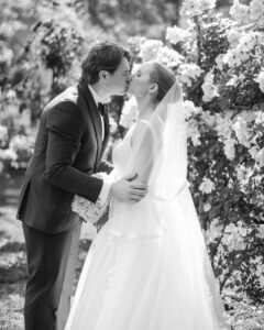A bride and groom kiss outdoors in front of blooming flowers at the Jasper Crane Rose Garden. The bride wears a gown and veil; the groom is in a suit. The black-and-white scene beautifully captures their wedding photos amidst the romantic garden setting.