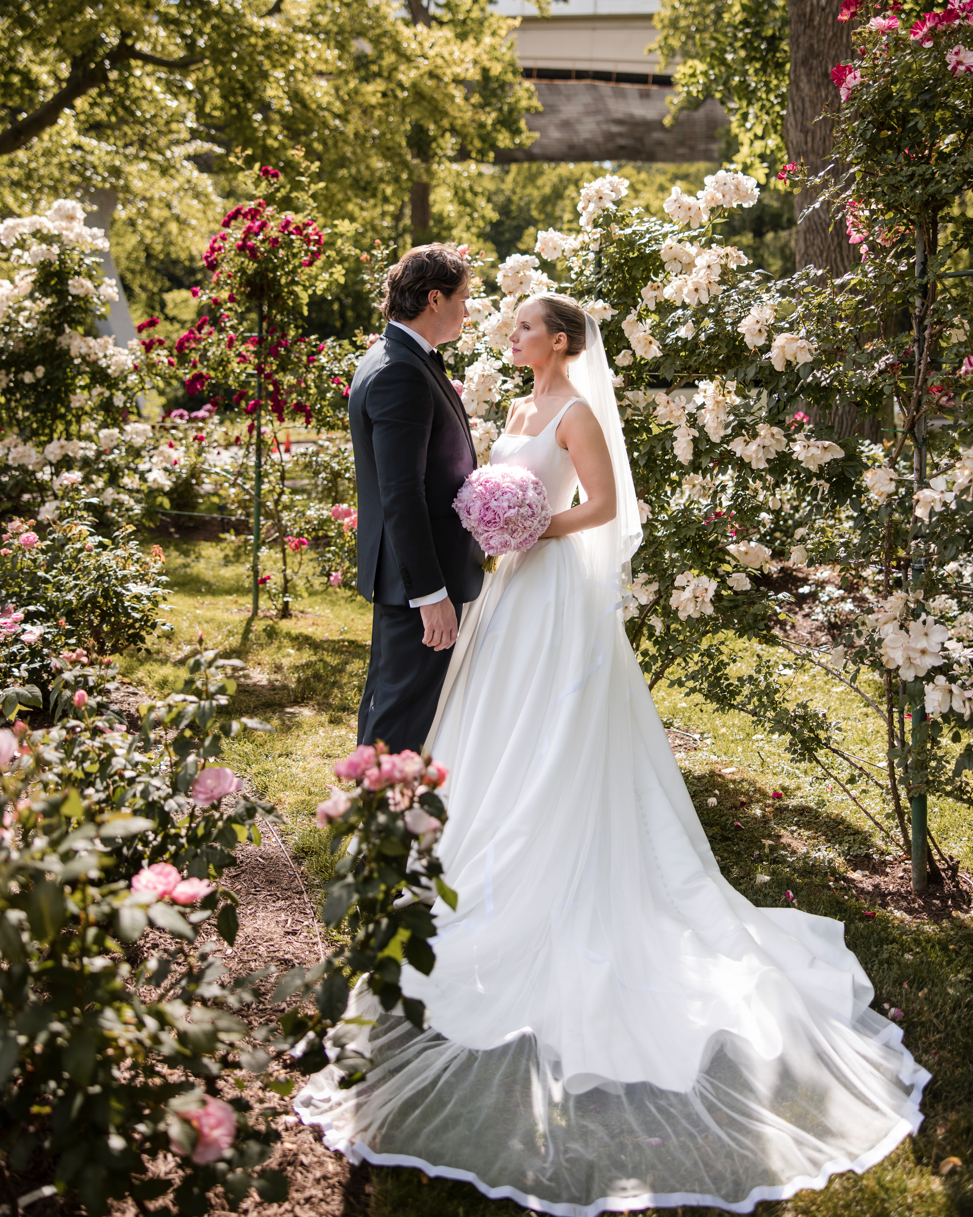 A bride and groom stand facing each other in a garden surrounded by blooming flowers, with the bride holding a bouquet and wearing a long white dress and veil—perfect for Wedding Photos at Jasper Crane Rose Garden.