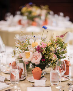 A round table set for the Longwood Gardens welcome party features a floral centerpiece, glasses of water, small terracotta pots, place cards, and neatly arranged tableware.