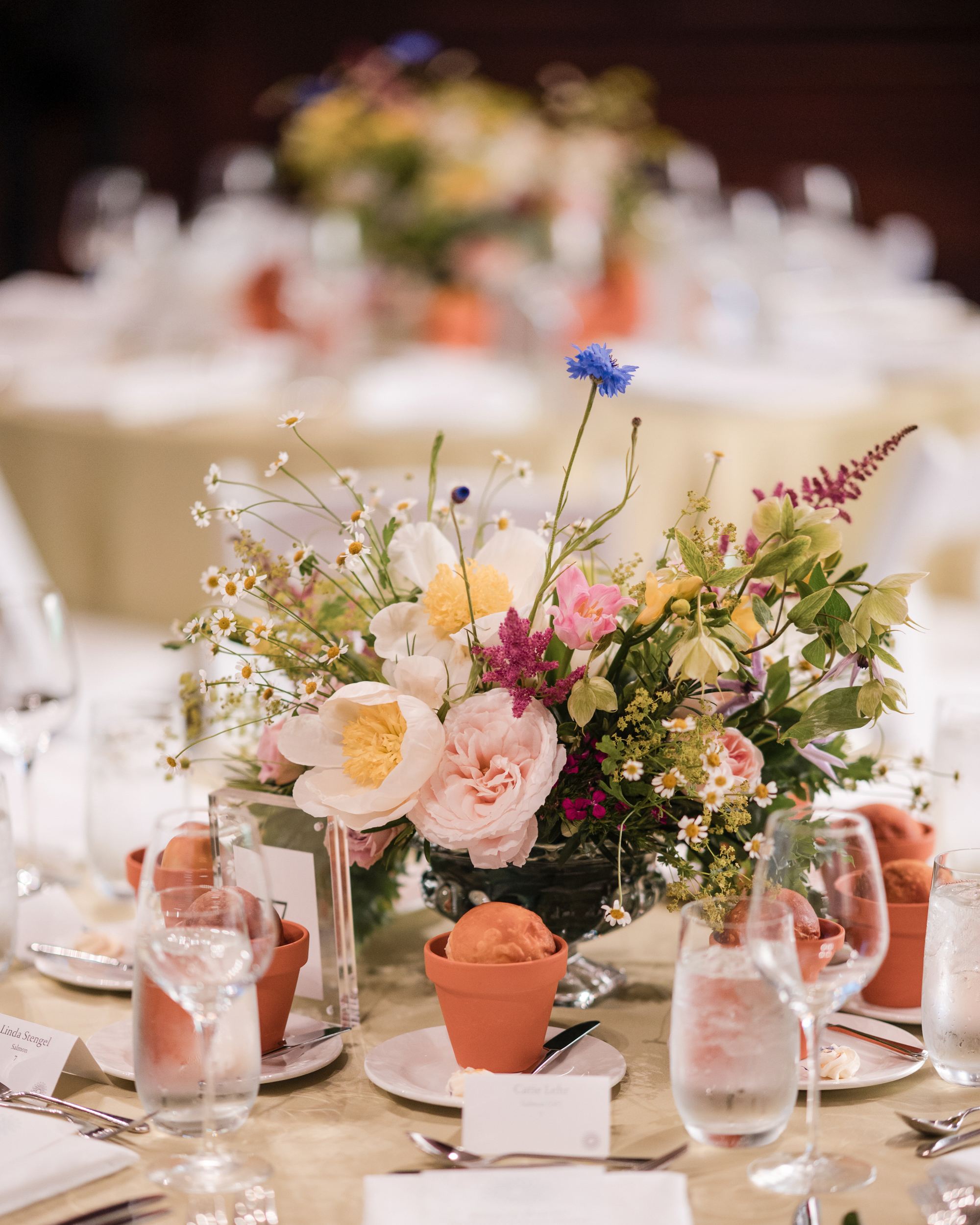 A round table set for the Longwood Gardens welcome party features a floral centerpiece, glasses of water, small terracotta pots, place cards, and neatly arranged tableware.
