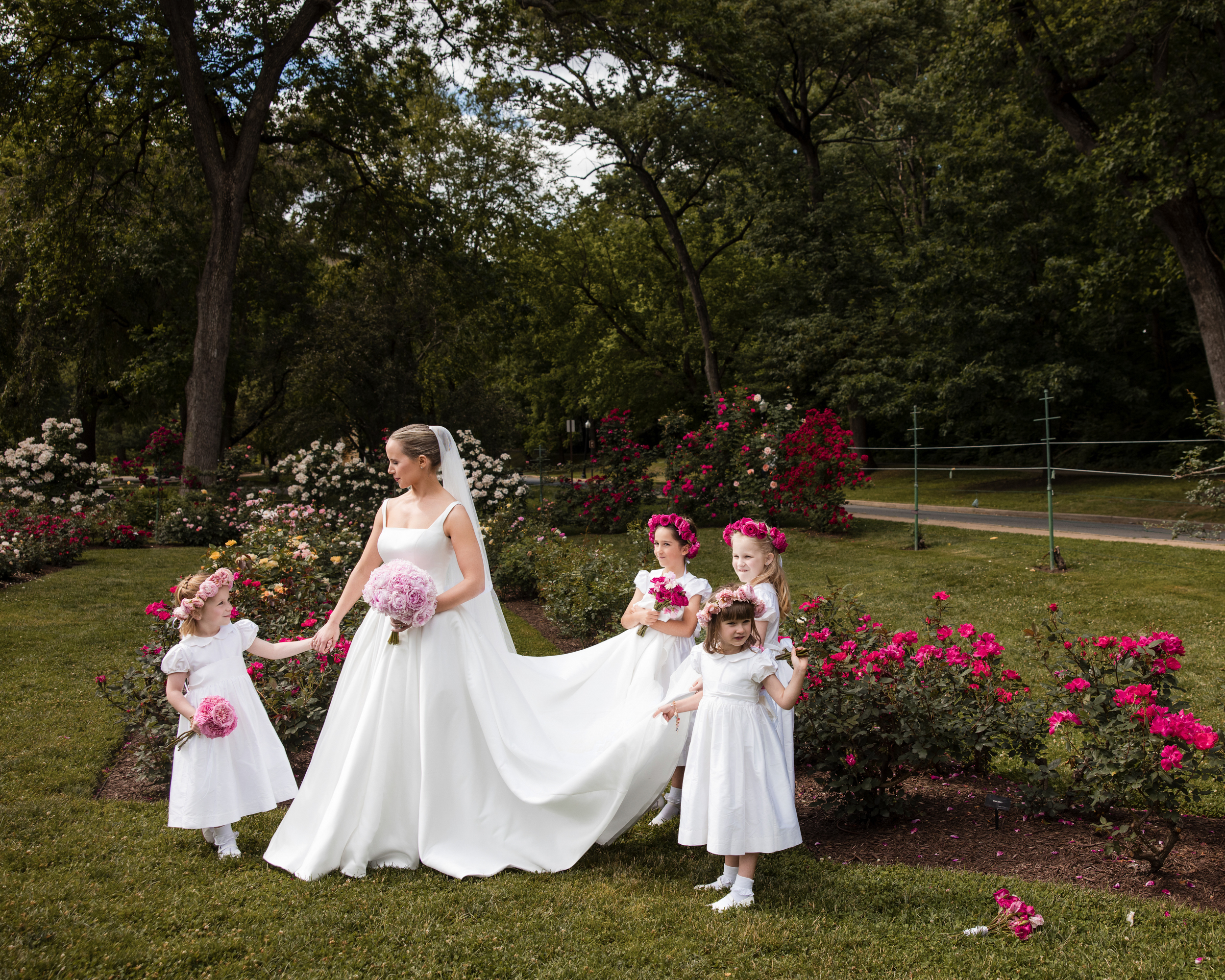 A bride in a white gown holds a bouquet and stands with four flower girls in white dresses, surrounded by blooming pink and red flowers—perfect for wedding photos at Jasper Crane Rose Garden. A bride in a white gown holds a bouquet and stands with four flower girls in white dresses, surrounded by blooming pink and red flowers—perfect for wedding photos at Jasper Crane Rose Garden.